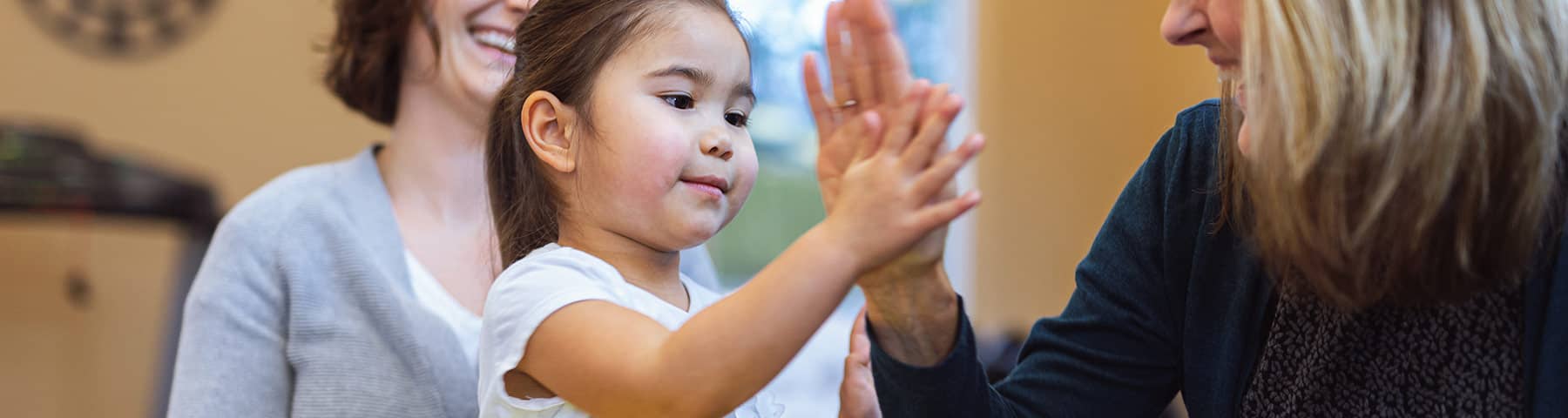 Young child smiling and giving a high-five to an adult, with a caregiver in the background, capturing a warm and collaborative moment in a supportive environment.