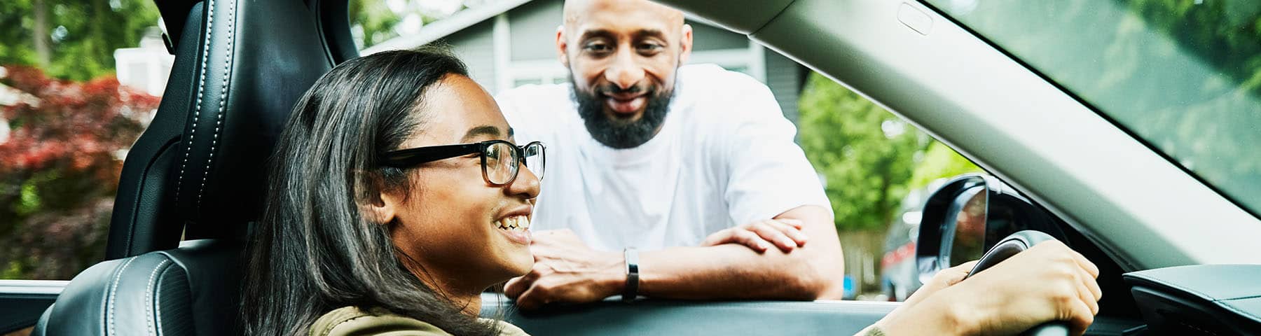 Teen driver smiling behind the wheel of a car while an adult leans on the window, offering guidance and encouragement in a safe and supportive setting.