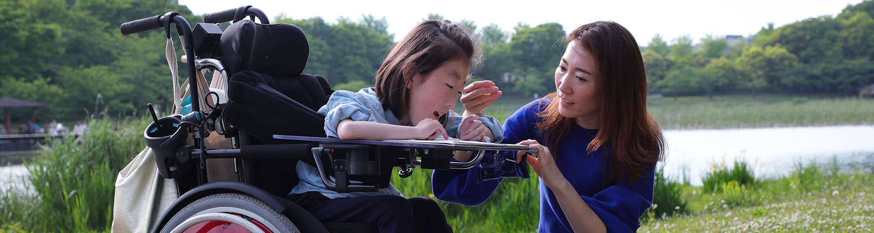 Child in a wheelchair receiving assistance outdoors by a caregiver near a scenic lake.