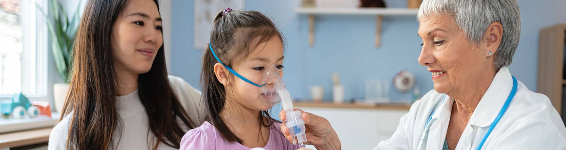 Child using a nebulizer mask with the assistance of a doctor, while a parent looks on.