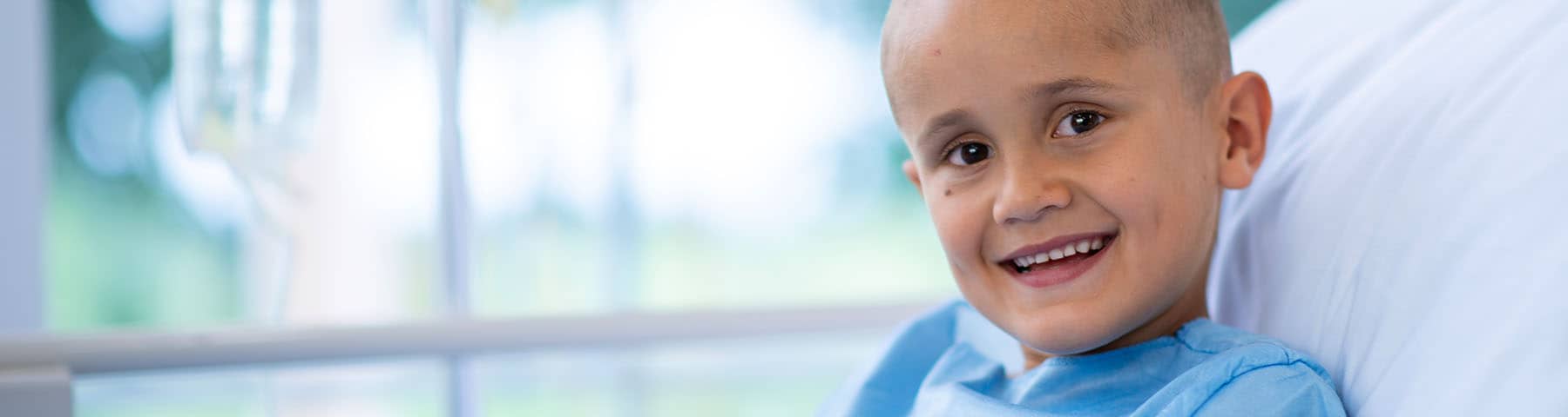 Smiling child in a hospital bed wearing a medical gown with an IV in the background.