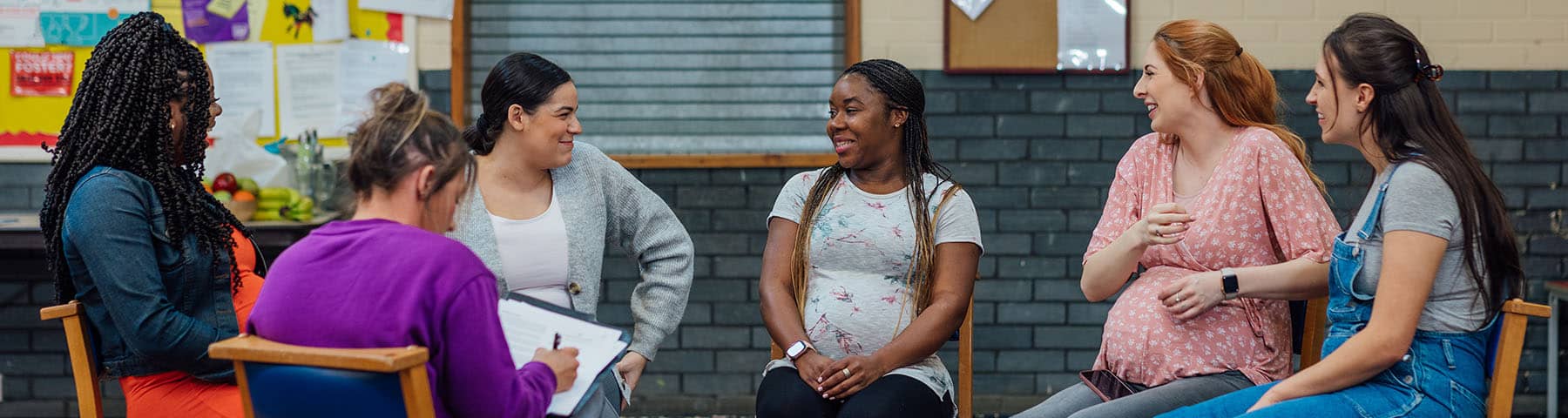 Group of diverse women, including expectant mothers, seated in a circle during a prenatal class, sharing smiles and conversation in a supportive and welcoming environment.