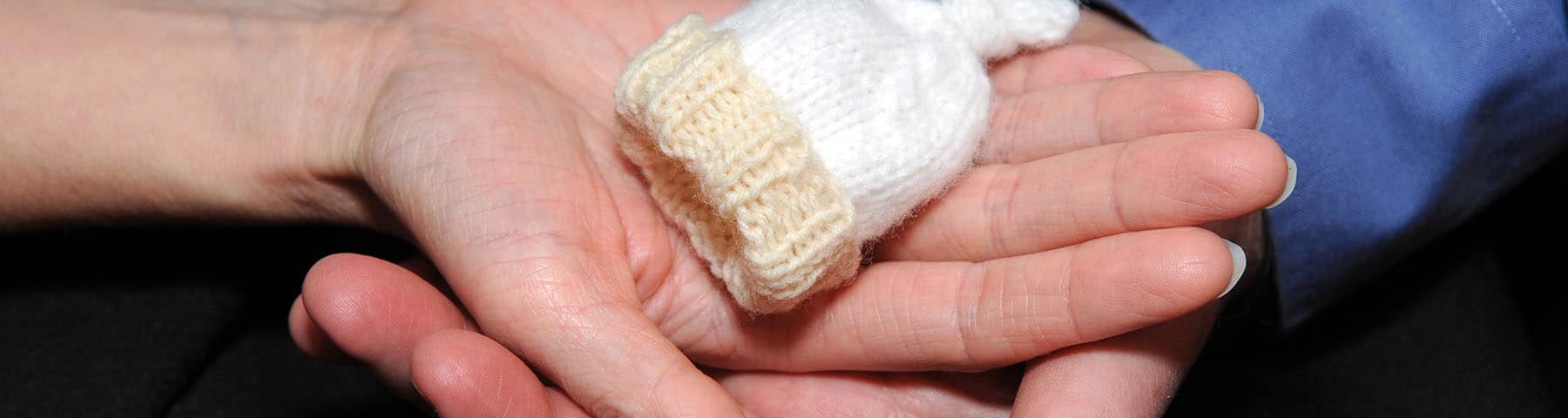 Close-up of an adult's hands gently cradling a tiny baby hand wearing a knitted mitten, symbolizing care, protection, and love.