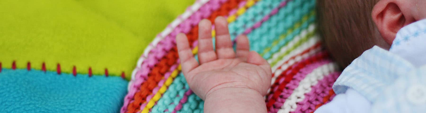 Close-up of a newborn's hand resting on a colorful knit blanket, symbolizing warmth, safety, and care.