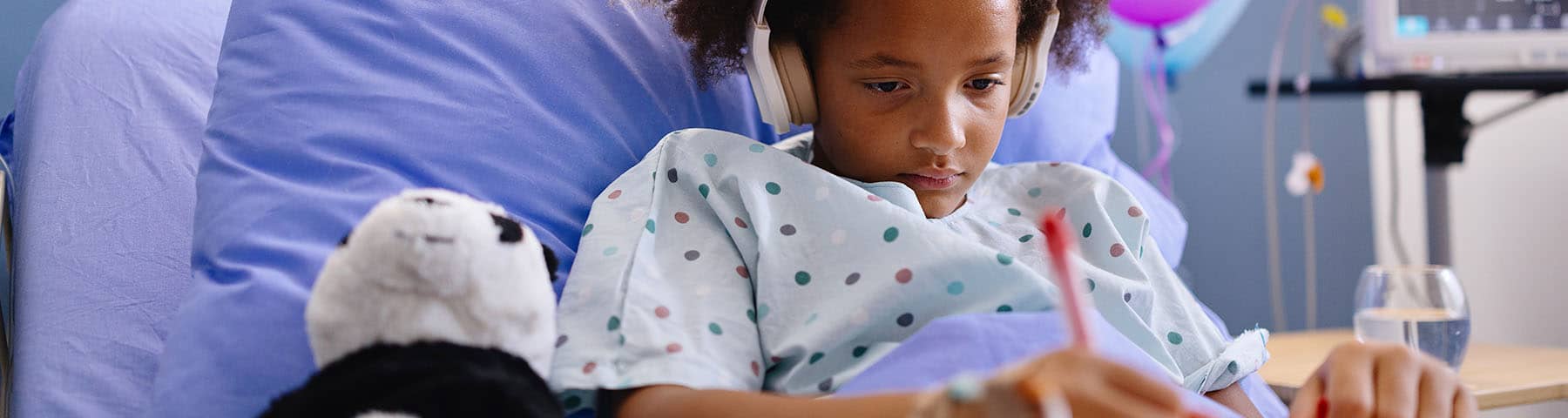 Young patient in a hospital bed, wearing headphones and focused on writing or drawing, with a stuffed animal beside them, creating a calming and engaging environment.