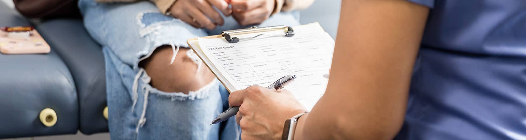 Close-up of a healthcare professional holding a clipboard with a patient chart, seated next to a patient wearing ripped jeans, creating a supportive and approachable clinical environment.