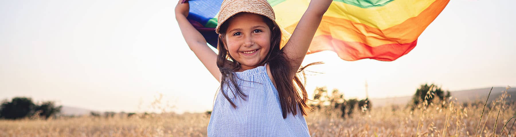 Smiling girl holding a rainbow flag in a sunny outdoor field.