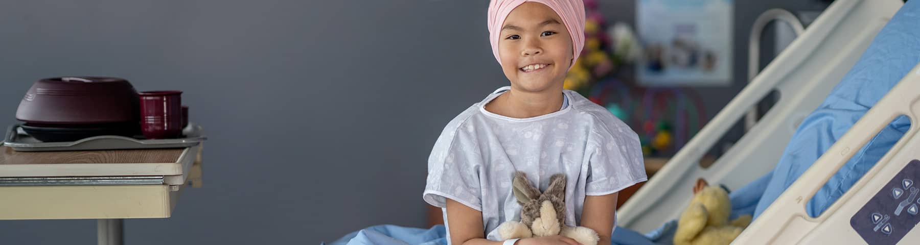 Young patient wearing a pink headscarf and hospital gown, sitting on a hospital bed and holding a stuffed rabbit, with a warm smile in a comforting medical setting.