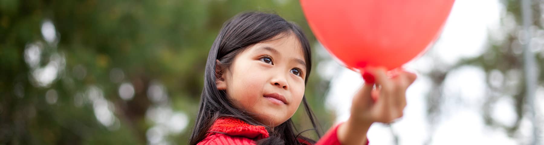 A young girl wearing a red sweater holds a red balloon outdoors, with blurred green foliage in the background.