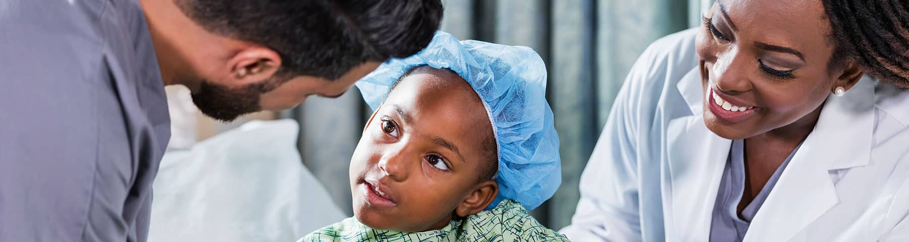 A young child wearing a blue surgical cap and hospital gown looks up attentively at a smiling doctor and a healthcare professional in a hospital setting.