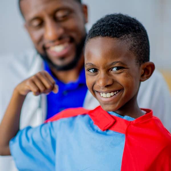 A smiling young boy flexing his muscle and wearing a red cape with his pediatrician smiling behind him.