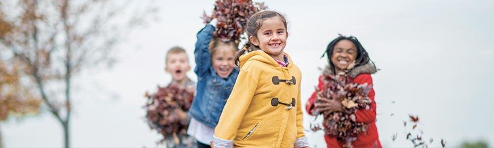 Children playing outdoors, smiling and tossing autumn leaves into the air.